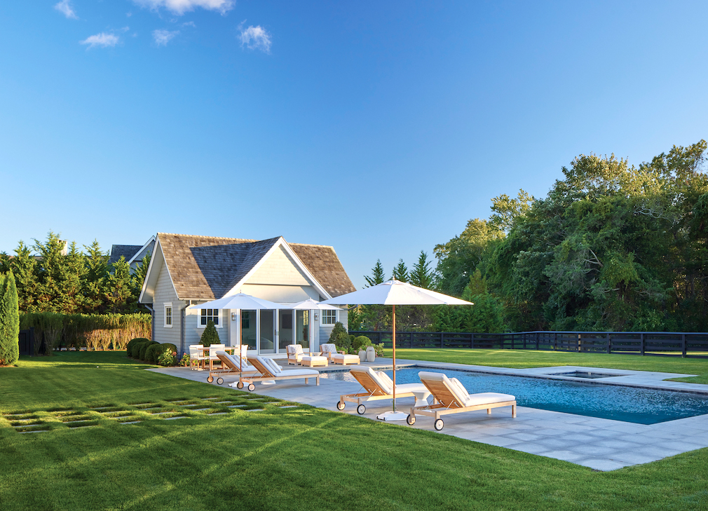 A pool and pool house with white lounge chairs and umbrella in the backyard of a Hamptons home
