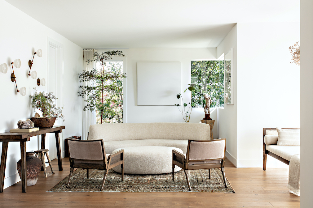 In the living room of this LA home a white sofa and ottoman were custom designed by Fogel Interiors in a boucle fabric from Una Malan and are positioned with antique side chairs found on 1stDibs. An antique rug from Woven and aged brass wall sconces by Apparatus.
